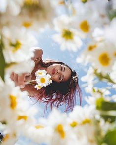 a woman holding a flower in her hand and looking up into the sky with daisies all around her