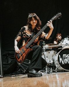 a woman sitting on the floor with a guitar in front of her and another drummer behind her