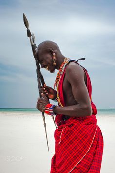 a man in red and white dress holding a spear on the beach with an ocean in the background