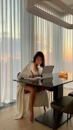 a woman sitting at a table with a laptop computer in her lap and writing on the paper