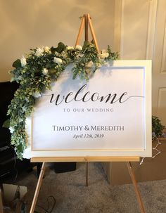 a welcome sign with flowers and greenery hanging from it's easel in front of a piano