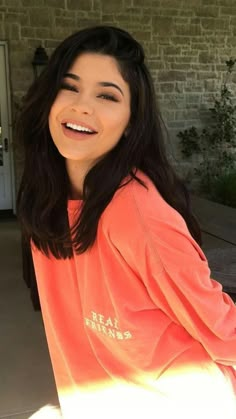 a woman with long dark hair wearing an orange shirt and smiling at the camera while sitting on a porch