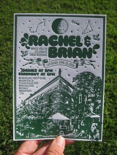 a hand holding up a poster for a concert in the grass with trees and buildings on it