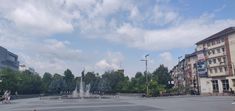 people are sitting on benches in the middle of a square with a fountain and trees