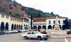 an empty parking lot with cars parked in front of buildings and trees on the hill