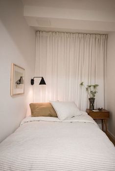 a white bed sitting under a window next to a wooden table with a lamp on it