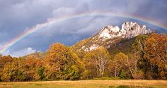 a rainbow in the sky over a field with trees and mountains behind it on a cloudy day