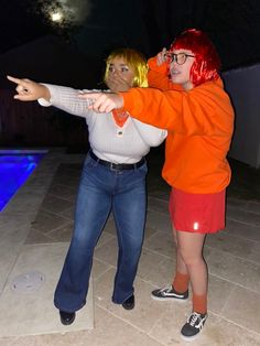 two women standing next to each other near a pool at night, one pointing her finger towards the camera