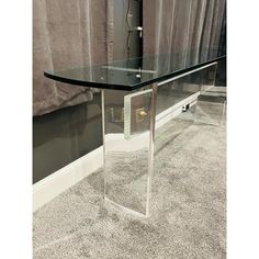 a glass and metal console table in the middle of a carpeted room with curtains behind it