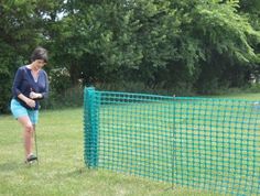 a woman standing next to a green fence with a ball in her hand and a golf club on the other side