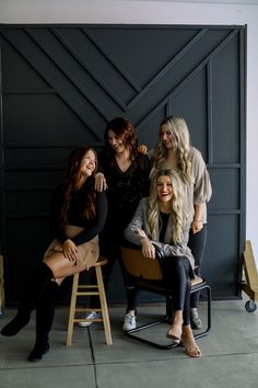 four women sitting on chairs posing for a photo in front of a garage door with their arms around each other