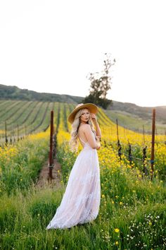 a woman in a long white dress and hat standing in a field with yellow flowers