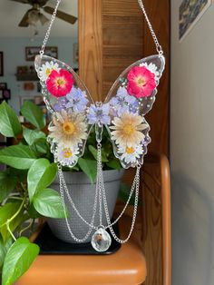 a butterfly shaped necklace with flowers attached to it's wings on a table next to a potted plant