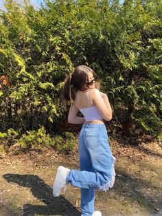 a woman in jeans and white top standing next to bushes with her hands behind her head