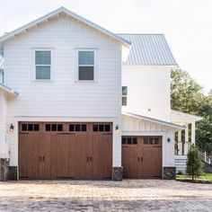 two brown garage doors are open in front of a white house with brick driveway and trees