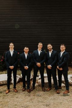 a group of men standing next to each other in front of a black wall wearing suits and ties