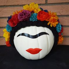 a white pumpkin decorated with colorful flowers and a face painted on the front, sitting in front of a brick wall