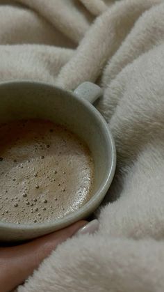 a person is holding a cup of hot chocolate in their hand on a white blanket