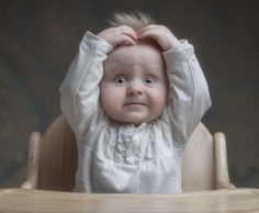 a baby sitting in a wooden highchair with its hands on the head and eyes open