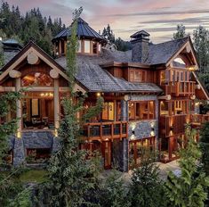 a large wooden house surrounded by trees in the woods at dusk with lights on it's windows