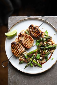 a white plate topped with grilled meat and asparagus next to a lime wedge