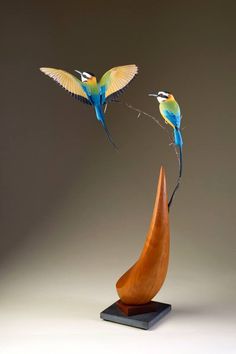 two blue and yellow birds are flying over a wooden sculpture on a black base with a white background