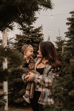 a woman holding a baby in her arms and looking at christmas trees with lights strung above them