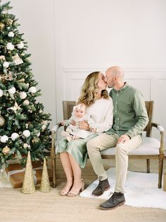 a man and woman are sitting next to a christmas tree with a baby in their lap