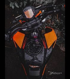 an orange and black motorcycle parked in front of some bushes with snow on the seat