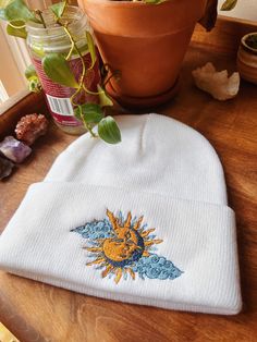 a sun and moon embroidered on a white beanie sits next to a potted plant