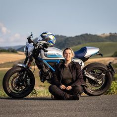 a woman sitting in front of a motorcycle on the side of the road with her legs crossed