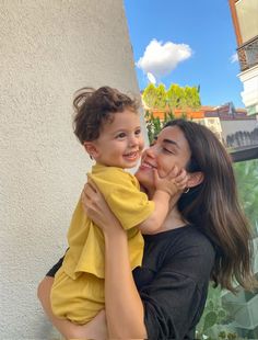 a woman holding a small child in her arms and smiling at the camera while standing next to a building