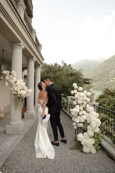 a bride and groom kissing in front of an elegant house with white flowers on the balcony