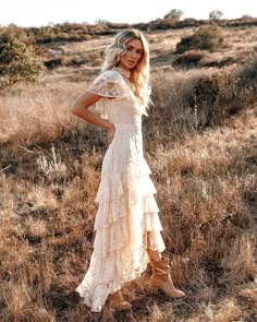 a woman standing in a field wearing a white dress and cowboy boots with her hands on her hips