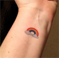 a small rainbow tattoo on the wrist with clouds and a rainbow in the sky above it