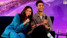 a man and woman sitting next to each other in front of a purple background holding microphones