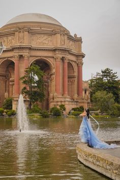 a woman in a blue dress standing next to a fountain with a bird flying over it