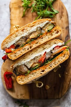 two sandwiches are on a cutting board with peppers and herbs in the background, ready to be eaten