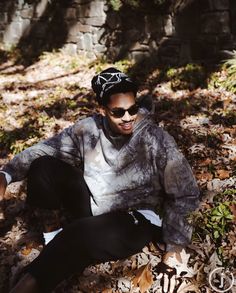 a young man sitting on the ground wearing sunglasses and a hat with his hands in his pockets