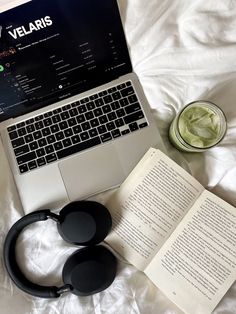 an open laptop computer sitting on top of a bed next to headphones and a book