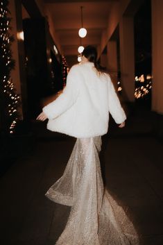 a woman in a white fur coat walking down a long hallway with lights on the walls