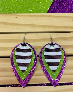 two pairs of purple and green glittered earrings on top of a wooden bench with grass in the background