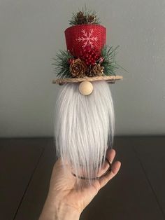 a hand holding up a white wig with pine cones on it and a red santa hat