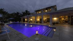 a large swimming pool in front of a house at night with lights on and chairs around it