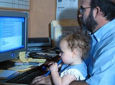 a man sitting in front of a computer monitor with a baby on his lap looking at the screen