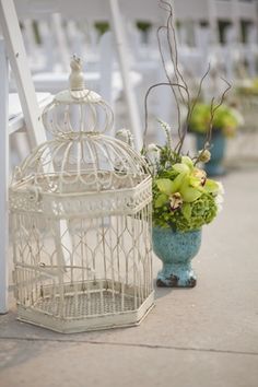 a white birdcage with flowers in it next to a blue vase filled with greenery