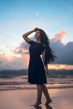 a woman standing on top of a sandy beach next to the ocean at sunset with her hair blowing in the wind