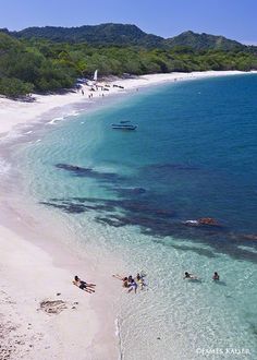 people are swimming in the clear blue water on a beach with white sand and green trees