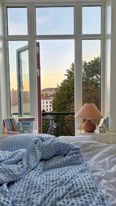 an unmade bed in front of two large windows with the view of a city