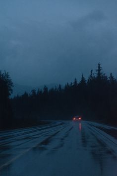 cars driving down a wet road with trees in the background at night, on a rainy day